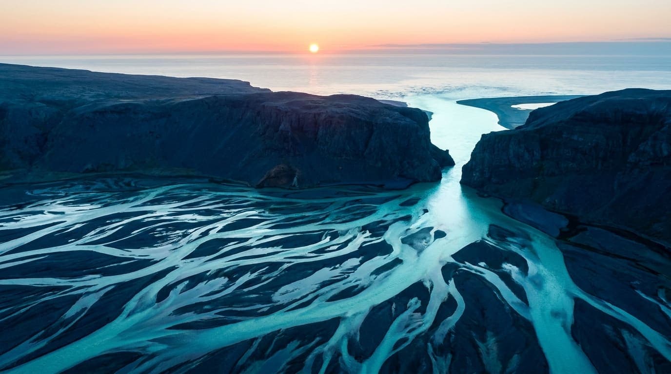 Aerial photograph of a braided river delta at sunrise, dozens of luminous channels converging through a single narrow gap between dark headlands.
